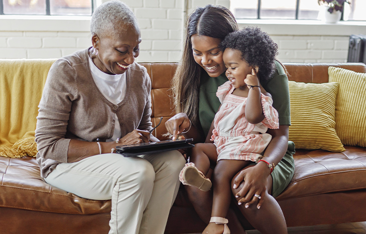 Trois générations (une jeune fille, sa mère et sa grand-mère) sourient sur un canapé en regardant un appareil électronique.